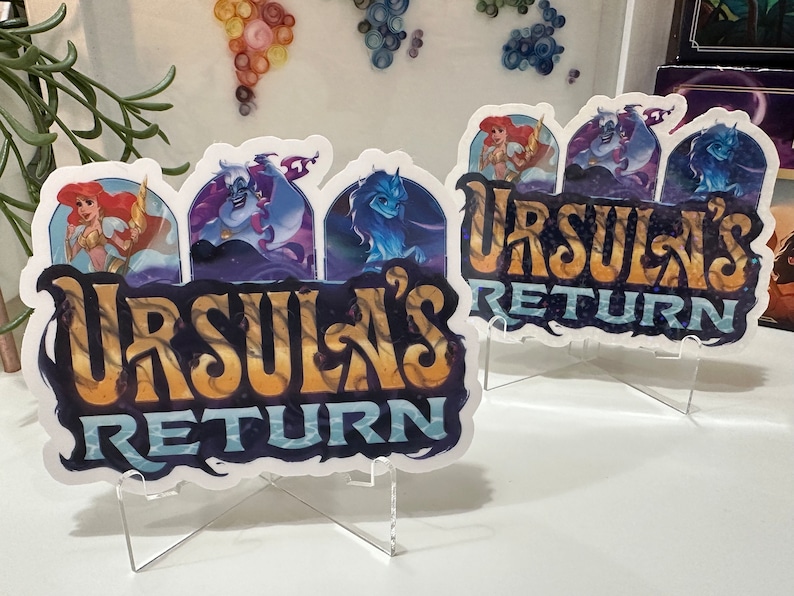 K&ouml;nnte beinhalten: Zwei wei&szlig;e Aufkleber mit einem blauen, violetten und goldenen Design. Die Aufkleber zeigen den Text "Ursula's Return" und Bilder von Ursula, einer Meerjungfrau und einer Meereskreatur. Die Aufkleber befinden sich auf klaren Acrylst&auml;ndern.