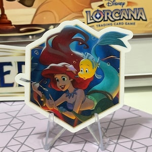 Puede incluir: Una pegatina hexagonal con Ariel de La Sirenita de Disney, con Flounder. La pegatina tiene un borde blanco y se muestra en un soporte de plástico transparente. El fondo muestra el logotipo del juego de cartas coleccionables Disney Lorcana.