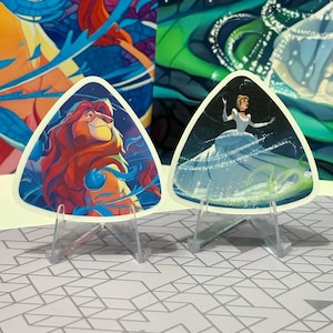 May include: Two triangular stickers on clear stands. One features Simba from The Lion King, the other Cinderella. The Simba sticker has a colorful design with orange, red, and blue hues. The Cinderella sticker shows her in a blue gown.