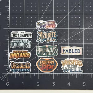 May include: A collection of colorful stickers with various fantasy-themed text labels, including "Shimmering Skies," "Azurite Sea," and "Ursula's Return." The stickers are arranged on a black grid surface.