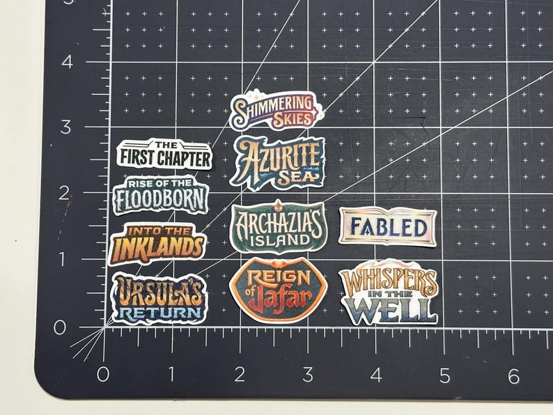 May include: A collection of colorful stickers with various fantasy-themed text labels, including "Shimmering Skies," "Azurite Sea," and "Ursula's Return." The stickers are arranged on a black grid surface.