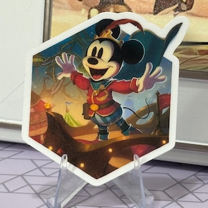Puede incluir: Una colorida pegatina con Mickey Mouse con un atuendo de circo, con una chaqueta roja, pantalones azules y un sombrero con plumas. El fondo muestra una carpa de circo y banderas. La pegatina se muestra en un soporte de plástico transparente.