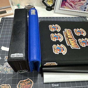 May include: A collection of card storage accessories, including a black box with "THE FIRST CHAPTER" text, a blue binder, and a black binder with "INTO THE INKLANDS" stickers. Various stickers are also visible.