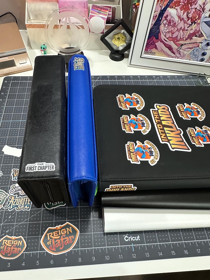 May include: A collection of card storage accessories, including a black box with "THE FIRST CHAPTER" text, a blue binder, and a black binder with "INTO THE INKLANDS" stickers. Various stickers are also visible.