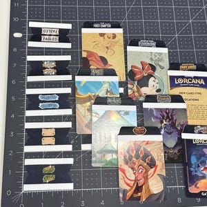 May include: A collection of Disney Lorcana trading card game dividers featuring various character and set illustrations including The First Chapter, Rise of the Floodborn, and Ursula's Return, arranged on a black cutting mat with white grid lines.