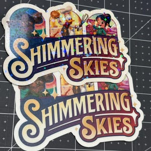 K&ouml;nnte beinhalten: Zwei Aufkleber mit glitzerndem, irisierendem Finish. Die Aufkleber zeigen eine farbenfrohe Illustration von Figuren aus dem Disney-Film "Encanto" und den Text "Shimmering Skies".