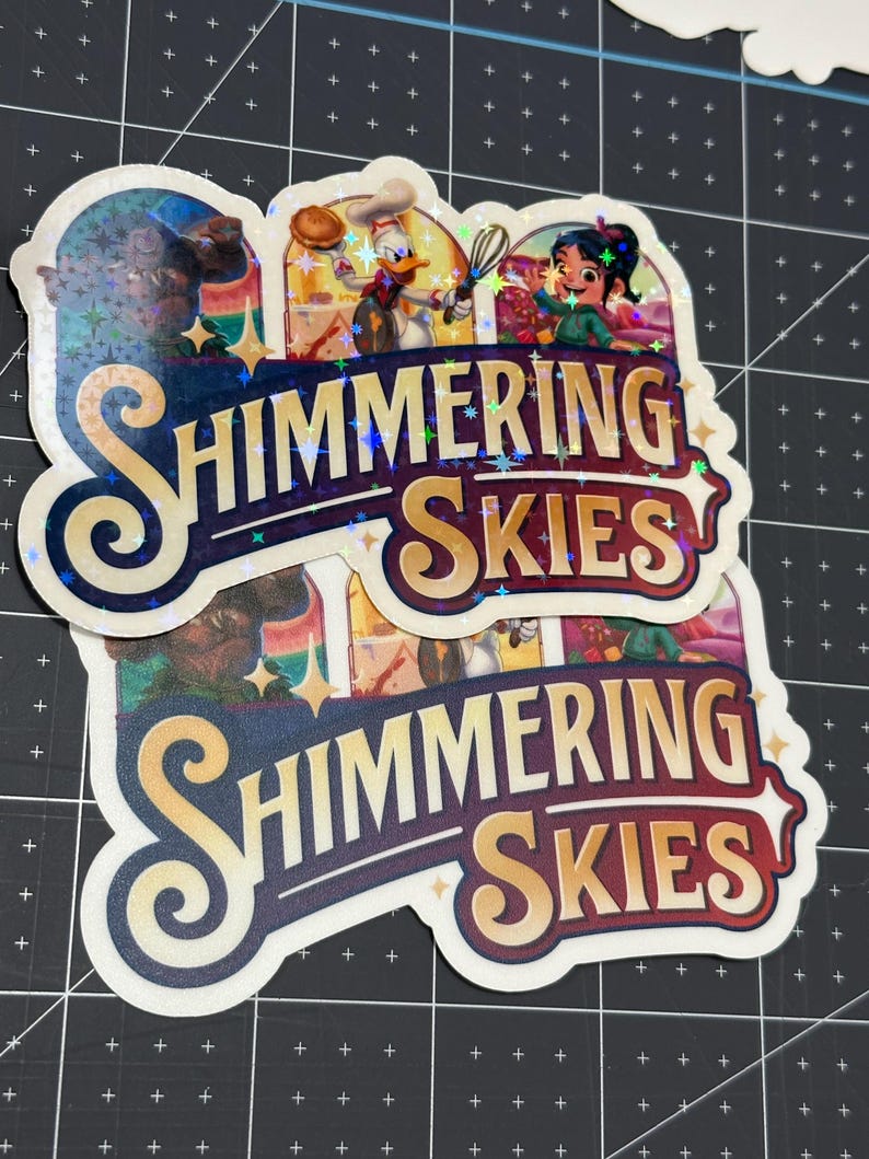 K&ouml;nnte beinhalten: Zwei Aufkleber mit glitzerndem, irisierendem Finish. Die Aufkleber zeigen eine farbenfrohe Illustration von Figuren aus dem Disney-Film "Encanto" und den Text "Shimmering Skies".