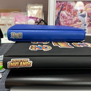 May include: A blue Azurite Sea binder sits atop a black binder with stickers and the text "Into the Inklands". The binders are stacked on top of a black and white surface.