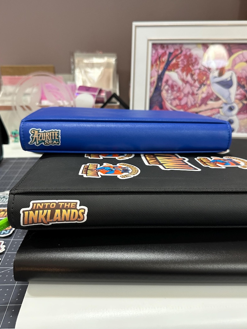 May include: A blue Azurite Sea binder sits atop a black binder with stickers and the text "Into the Inklands". The binders are stacked on top of a black and white surface.