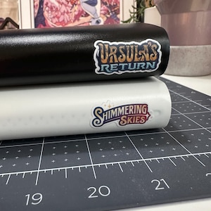 May include: Black and white cylindrical objects with stickers. The black object has a sticker that reads "URSULA'S RETURN" in a stylized font. The white object has a sticker that reads "SHIMMERING SKIES". The objects are on a black cutting mat.
