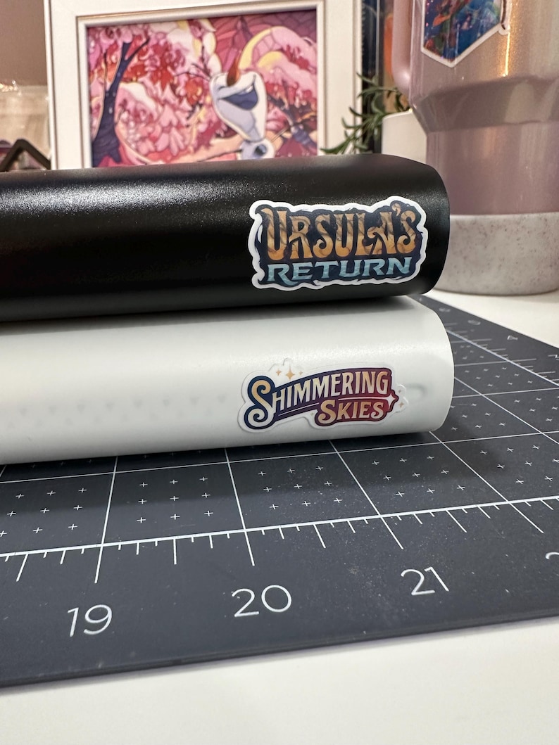 May include: Black and white cylindrical objects with stickers. The black object has a sticker that reads "URSULA'S RETURN" in a stylized font. The white object has a sticker that reads "SHIMMERING SKIES". The objects are on a black cutting mat.