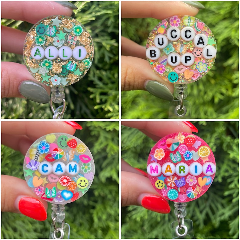 May include: Four circular badge reels featuring resin designs with embedded colorful charms and letter beads. The designs include names like ALLI, BUCCAL UP, CAM, and MARIA, surrounded by various shapes like flowers, hearts, and smiley faces.