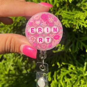 May include: A circular badge reel featuring a clear resin base filled with pink glitter, small heart, star, flower, smiley face, and lip-shaped confetti. The center displays white beads with pink letters spelling ERIN and RT in two rows.