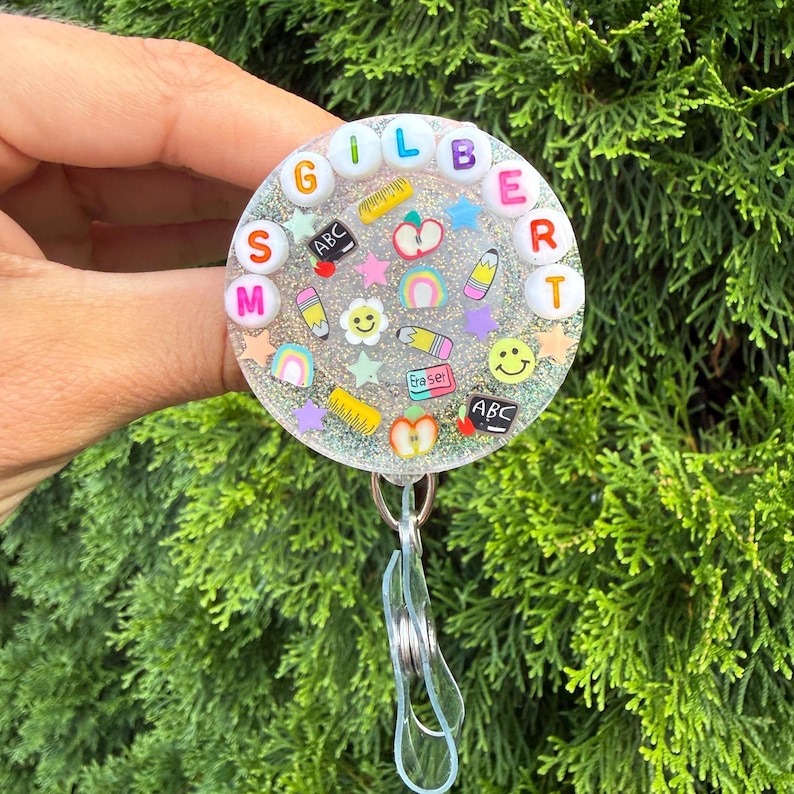 May include: A circular badge reel with a glittery resin finish featuring the text MS GILBERT spelled out in colorful letter beads. The surface is decorated with small school-themed charms including pencils, apples, rulers, and smiley faces.