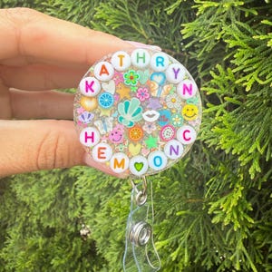 May include: A circular badge reel featuring a clear resin dome filled with colorful miniature charms including flowers, hearts, stars, and fruit slices. The name KATHRYN is spelled out in white alphabet beads around the top edge of the circle.