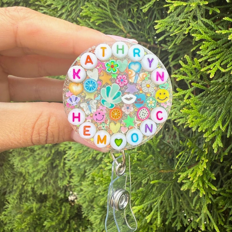 May include: A circular badge reel featuring a clear resin dome filled with colorful miniature charms including flowers, hearts, stars, and fruit slices. The name KATHRYN is spelled out in white alphabet beads around the top edge of the circle.
