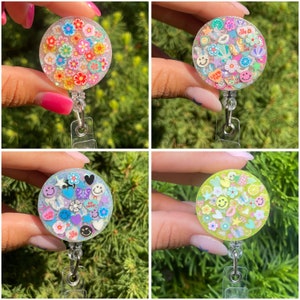 May include: A set of four circular badge reels featuring clear resin filled with colorful polymer clay slices. The designs include various floral patterns, smiley faces, hearts, stars, and text accents like 'love you', 'hug me', 'be mine', and 'so cute'.
