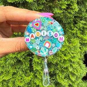 May include: A circular badge reel featuring a clear resin base filled with teal glitter and various colorful charms including hearts, rainbows, flowers, and smiley faces. The name MADISON is spelled out in white round beads across the center.