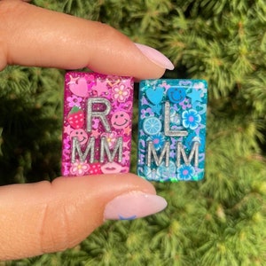 Puede incluir: Dos pequeños azulejos cuadrados de resina con purpurina y diseños coloridos. Los azulejos deletrean "RLMM" en letras plateadas con purpurina. Un azulejo es rosa con fresas, corazones y estrellas. El otro azulejo es azul con limones, flores y caras sonrientes.