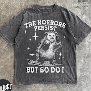 May include: Dark grey t-shirt with a distressed graphic of an opossum and the text "THE HORRORS PERSIST BUT SO DO I". The design includes stars and is from the brand "RETROCIDE STREETWEAR".