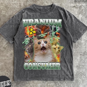 May include: A charcoal gray short-sleeve t-shirt featuring a graphic print of three cats against a fiery explosion background. The text reads URANIUM CONSUMER with a yellow radiation symbol and the phrase 20 BILLION CLORIC repeated twice.