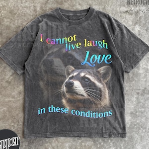 May include: A charcoal gray short-sleeve t-shirt featuring a graphic of a raccoon. The text reads I cannot live laugh Love in these conditions in a gradient of blue, pink, and yellow. The shirt has a faded, washed-out appearance throughout.