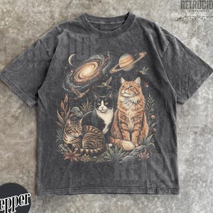 May include: Dark gray t-shirt featuring a colorful illustration of three cats with a galaxy and planets in the background. The cats are a mix of tabby, black and white, and orange. The shirt has a vintage, distressed look.
