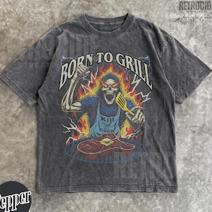 Puede incluir: Camiseta gris oscuro con un diseño de un esqueleto asando un filete, llamas y el texto "BORN TO GRILL". El esqueleto lleva un delantal azul con la frase "Kill the Cook". La camiseta tiene un aspecto vintage.