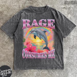May include: Dark gray t-shirt with a graphic of a dolphin leaping over a rainbow, with the words "RAGE CONSUMES ME" in pink. The design includes hibiscus flowers and a sunset scene.