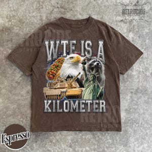 May include: Espresso-colored t-shirt with a graphic design featuring a hot dog, bald eagle, raccoon, tank, and the Statue of Liberty. The text "WTF IS A KILOMETER" is printed across the front. The shirt has a vintage, worn look.
