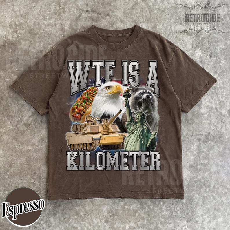 May include: Espresso-colored t-shirt with a graphic design featuring a hot dog, bald eagle, raccoon, tank, and the Statue of Liberty. The text "WTF IS A KILOMETER" is printed across the front. The shirt has a vintage, worn look.