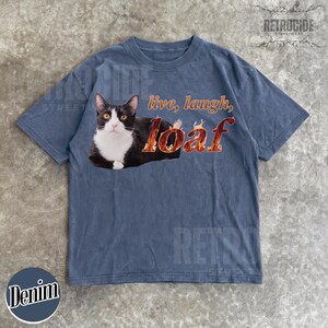 Puede incluir: Una camiseta de manga corta en color azul deste&ntilde;ido con un gr&aacute;fico de un gato blanco y negro tumbado. El texto "live, laugh, loaf" est&aacute; impreso en llamas naranjas y amarillas junto al gato. La camiseta tiene cuello redondo y corte holgado.