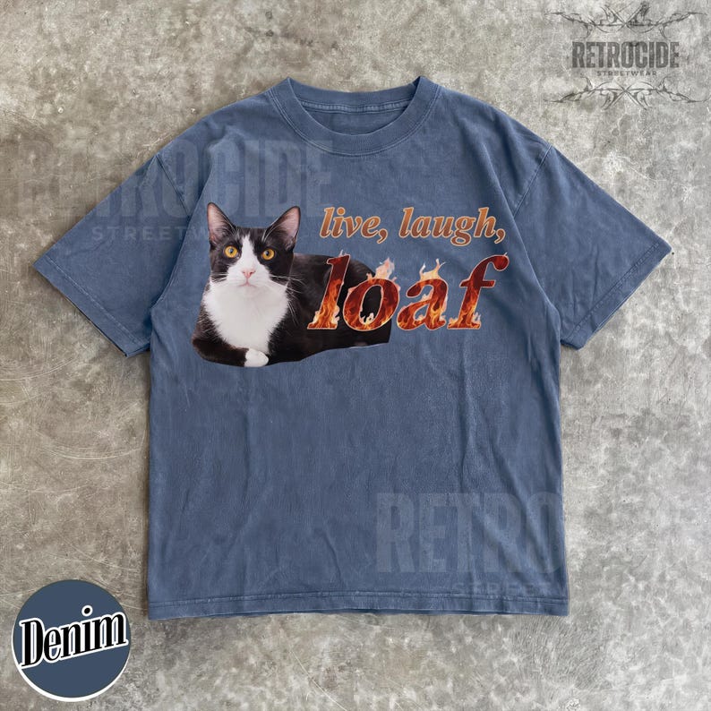 Puede incluir: Una camiseta de manga corta en color azul deste&ntilde;ido con un gr&aacute;fico de un gato blanco y negro tumbado. El texto "live, laugh, loaf" est&aacute; impreso en llamas naranjas y amarillas junto al gato. La camiseta tiene cuello redondo y corte holgado.