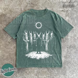 May include: Light green t-shirt with a white graphic of figures with horned masks and a sun-like symbol. The shirt has a crew neck and short sleeves. The text "RETROCIDE STREETWEAR" is visible in the upper right corner.