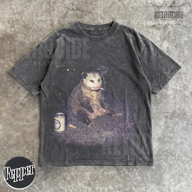 May include: Dark gray t-shirt featuring a graphic of an opossum smoking a cigarette, with a can and a cigarette butt. The shirt has a vintage, distressed look. The brand name "RETROCIDE STREETWEAR" is printed on the top right.