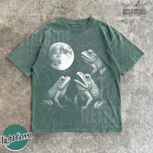 May include: Light green t-shirt featuring a detailed illustration of three frogs under a full moon. The frogs are in a circle, with one frog's mouth open. The shirt has the text "Light Green" in a circle at the bottom.