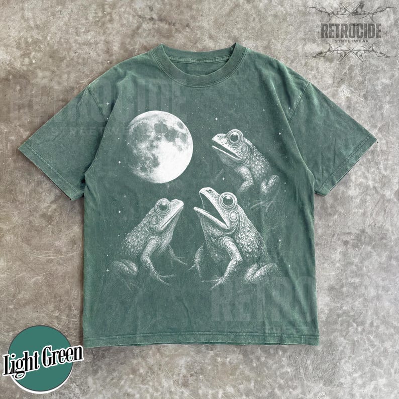 May include: Light green t-shirt featuring a detailed illustration of three frogs under a full moon. The frogs are in a circle, with one frog's mouth open. The shirt has the text "Light Green" in a circle at the bottom.
