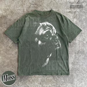 May include: An olive green short-sleeve t-shirt featuring a large, detailed white graphic illustration of a dog's face on the front. The design shows the side profile of the dog with visible fur texture and shading. The shirt has a crew neckline.