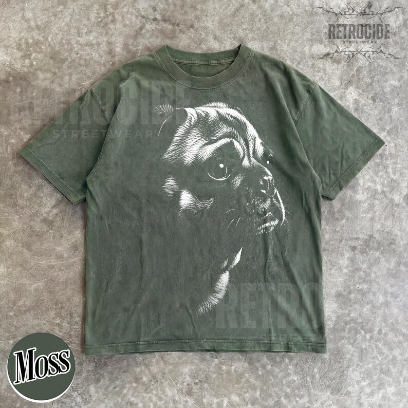May include: An olive green short-sleeve t-shirt featuring a large, detailed white graphic illustration of a dog's face on the front. The design shows the side profile of the dog with visible fur texture and shading. The shirt has a crew neckline.