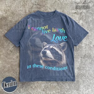 Puede incluir: Una camiseta gris carb&oacute;n de manga corta con un gr&aacute;fico de un mapache. El texto "I cannot live laugh Love in these conditions" aparece en una fuente de degradado multicolor. El dise&ntilde;o incluye una imagen grande y desvanecida de un mapache.