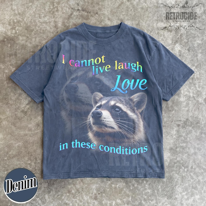 Puede incluir: Una camiseta gris carb&oacute;n de manga corta con un gr&aacute;fico de un mapache. El texto "I cannot live laugh Love in these conditions" aparece en una fuente de degradado multicolor. El dise&ntilde;o incluye una imagen grande y desvanecida de un mapache.