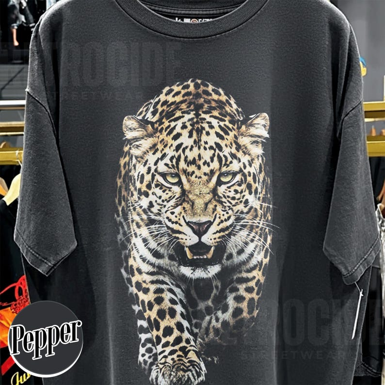 May include: Black t-shirt featuring a detailed leopard graphic. The leopard is depicted in shades of brown, tan, and black, with a fierce expression. The shirt has a vintage, distressed look. The text "ROCK" is visible at the top of the shirt.