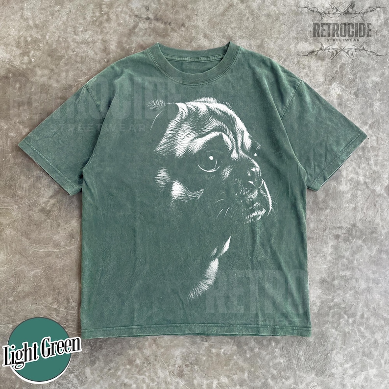 May include: A short-sleeve t-shirt in a muted sage green color featuring a large, detailed white graphic print of a dog's face in profile on the front. The shirt has a crew neckline and a standard fit, with the text RETROCIDE visible in the corner.