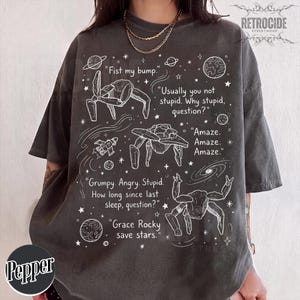 Könnte beinhalten: Ein anthrazitfarbenes Kurzarm-T-Shirt mit weißen Strichzeichnungen von Weltraumkreaturen, Planeten und Sternen. Die Grafik enthält den Text: "Fist my bump," "Usually you not stupid. Why stupid, question?," "Amaze. Amaze. Amaze.," "Grumpy. Angry. Stupid. How long since last sleep, question?," und "Grace Rocky save stars."