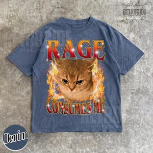Puede incluir: Una camiseta de manga corta en azul deste&ntilde;ido con un estampado gr&aacute;fico de una cara de gato rodeada de llamas naranjas y amarillas. El texto RAGE est&aacute; impreso en rojo sobre el gato y CONSUMES ME en rojo debajo, ambos con contornos blancos.