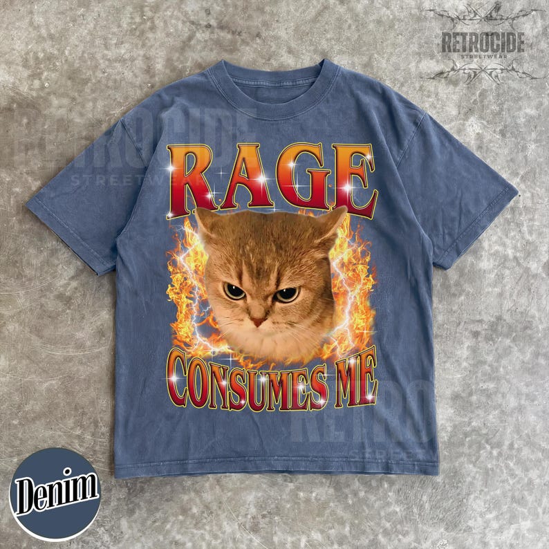 Puede incluir: Una camiseta de manga corta en azul deste&ntilde;ido con un estampado gr&aacute;fico de una cara de gato rodeada de llamas naranjas y amarillas. El texto RAGE est&aacute; impreso en rojo sobre el gato y CONSUMES ME en rojo debajo, ambos con contornos blancos.