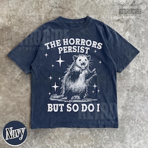 May include: Navy blue t-shirt with a distressed graphic of an opossum and the text "THE HORRORS PERSIST BUT SO DO I". The design includes stars and is printed in white. The shirt has a classic crew neck.