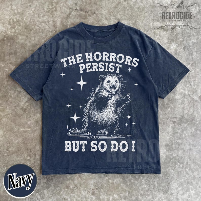 May include: Navy blue t-shirt with a distressed graphic of an opossum and the text "THE HORRORS PERSIST BUT SO DO I". The design includes stars and is printed in white. The shirt has a classic crew neck.