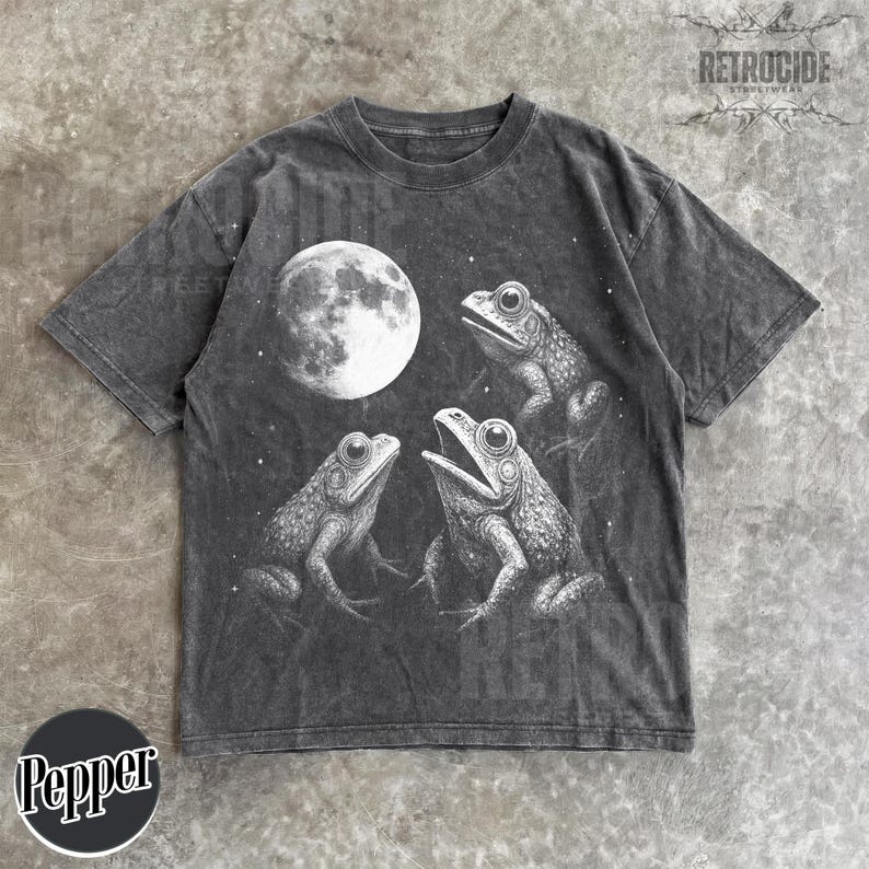 May include: Dark gray t-shirt featuring a detailed illustration of three frogs under a full moon. The design is in black and white, with the word "RETROCIDE" printed at the top. The shirt has a vintage, washed look.