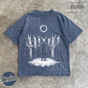 May include: Denim blue t-shirt featuring a white graphic of figures with horned masks and cloaks, under a sun-like symbol. The shirt has the text "RETROCIDE STREETWEAR" and "Denim" in a circle.